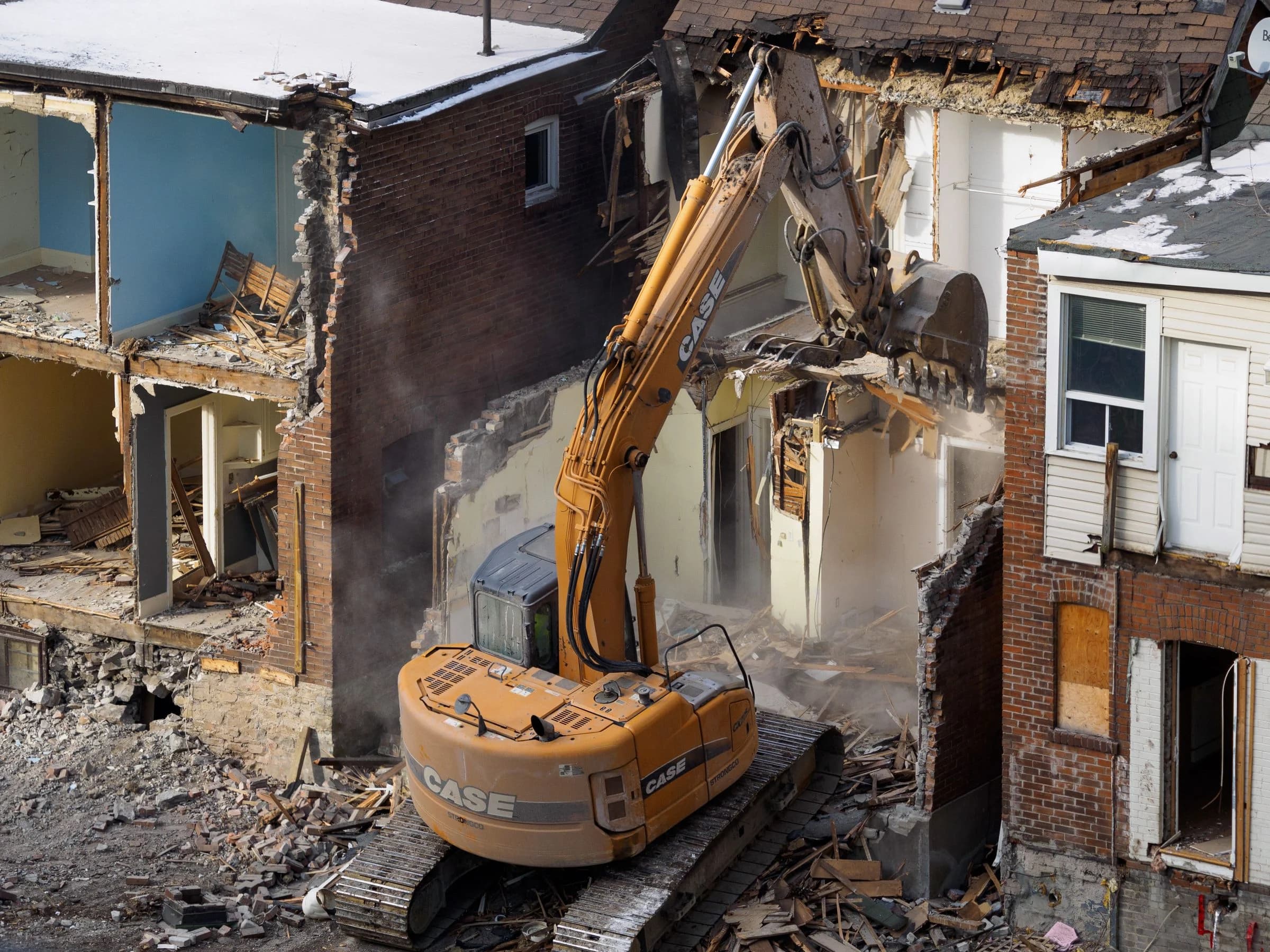 An excavator removing part of a brick building with exposed rooms and demolition debris